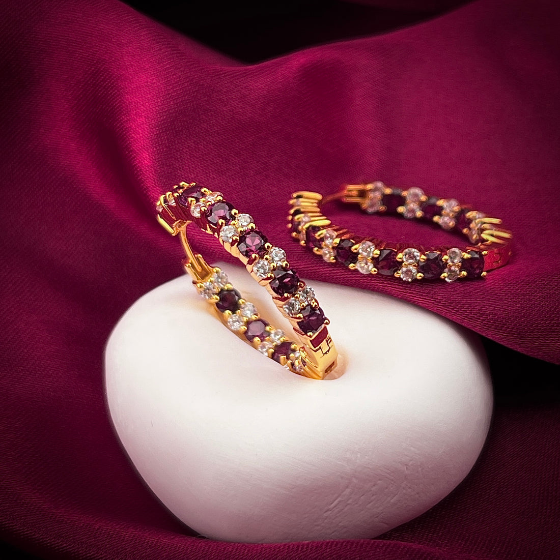Haspiera Rhodolite and cubic zirconia hoop earrings in gold setting on white stone with burgundy fabric