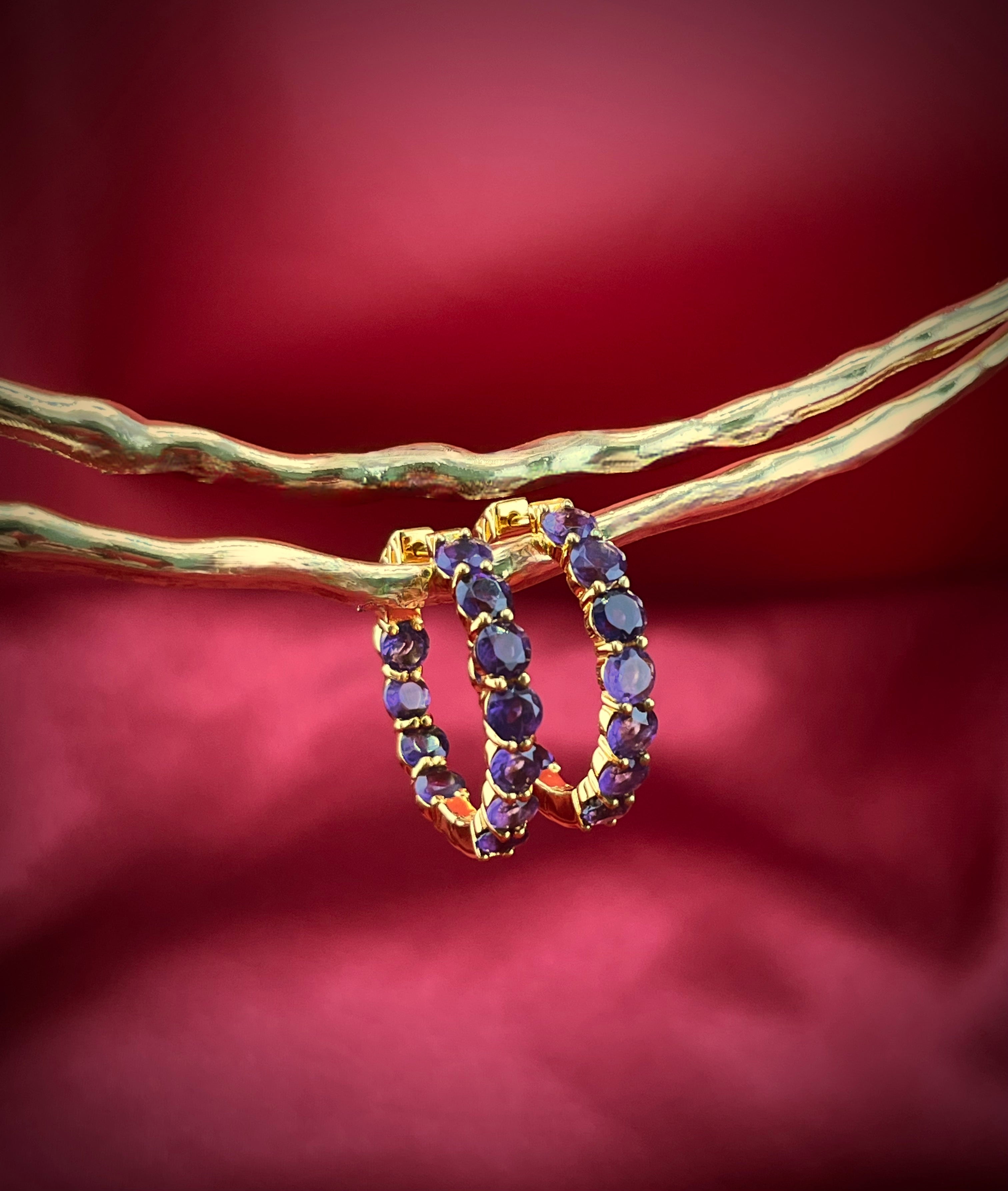 Haspiera amethyst hoop earrings with gold setting displayed on textured gold rods against a red background