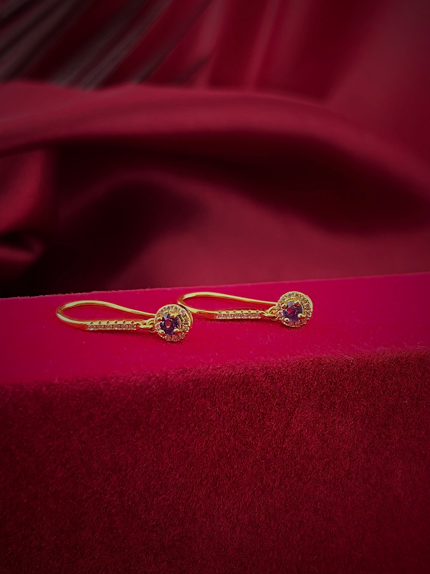 Haspiera gold gemstone violet drop earrings with intricate detailing on red velvet background