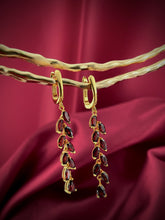 Haspiera red garnet dangle earrings with gold setting hanging on a textured branch against a red fabric background
