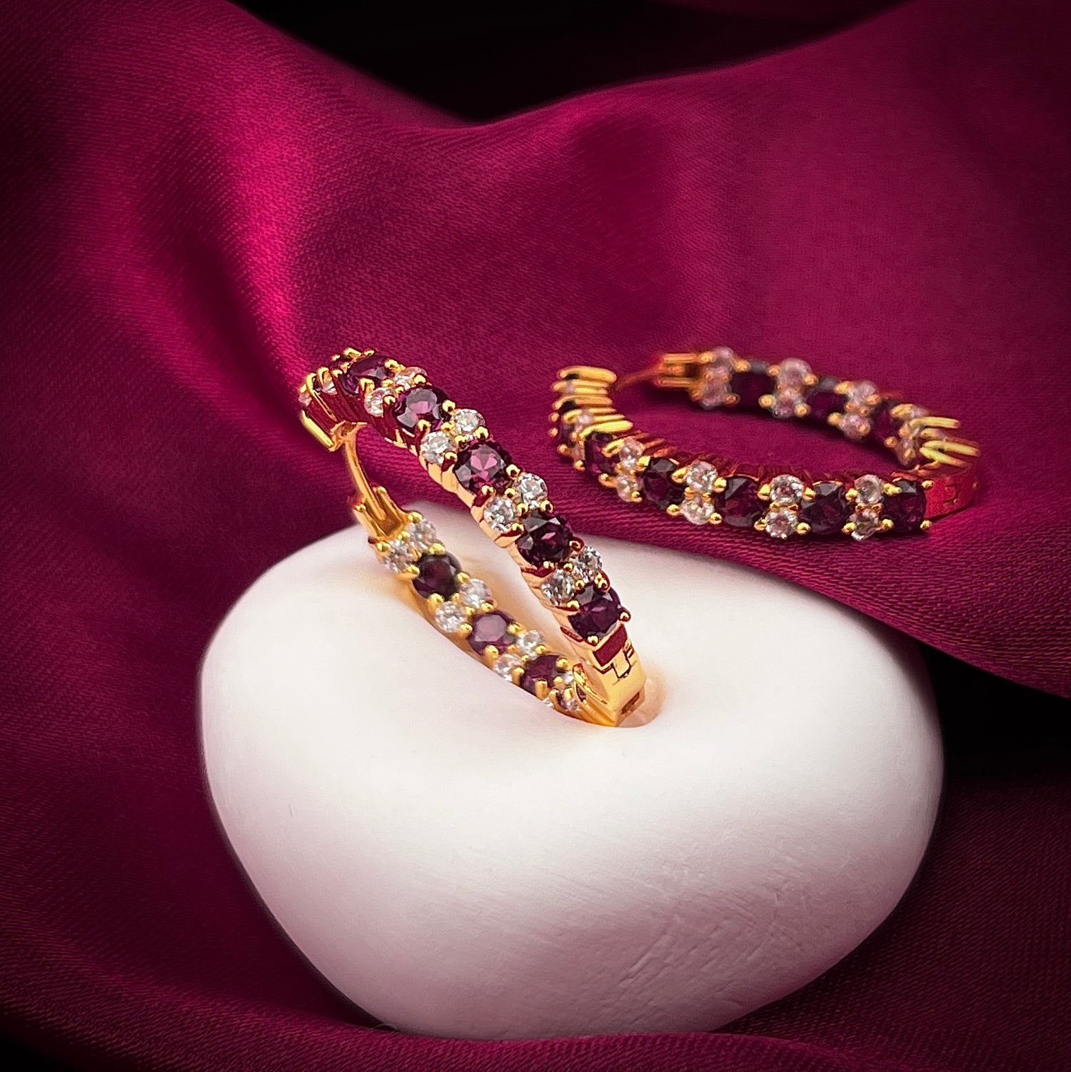 Haspiera Rhodolite and cubic zirconia hoop earrings in gold setting on white stone with burgundy fabric